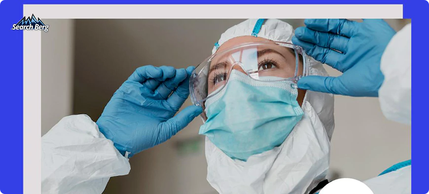 A healthcare worker dressed in full protective gear, including a white hazmat suit, blue gloves, a blue face mask, and clear protective goggles.